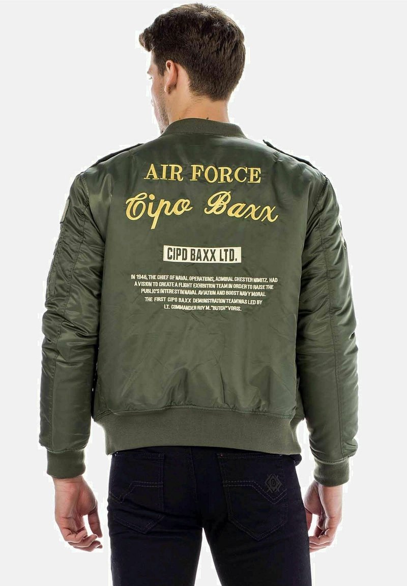 Olijfgroene bomberjack met gouden tekst op de achterkant; voorzien van ribgebreide manchetten en zoom; gepresteerde branding en historische tekstdetails.