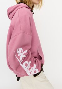 Felpa con cappuccio rosa in cotone con una grande stampa bianca "skidub" sulla manica. Presenta un cappuccio con coulisse e una vestibilità comoda.