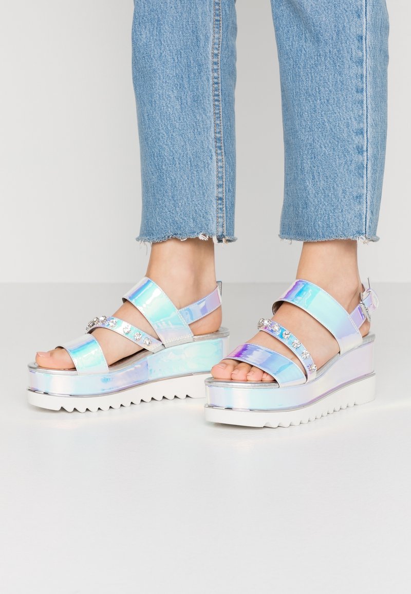 Pieds portant des sandales à plateforme iridescentes avec des brides en strass, associées à un jean denim bleu clair raccourci, sur un fond uni.