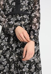 Czarna sukienka w kwiaty z przezroczystym materiałem, z szaro-białymi wzorami kwiatowymi, długimi rękawami, dopasowaną talią i satynowym paskiem w talii.