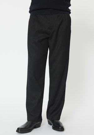 Mann trägt schwarze maßgeschneiderte Hose und schwarze Leder-Stiefeletten, steht vor einem schlichten, hellen Hintergrund.