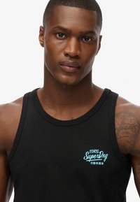 Top in cotone nero con logo "SuperDry" in teal e testo giapponese. Il design presenta un collo rotondo e spalline larghe.