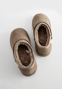 Pantoufles en daim marron avec doublure en peluche, bout arrondi et détail de fermeture éclair sur le côté. Texture douce et design simple, adaptées pour une utilisation intérieure.