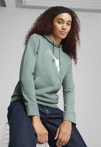 Felpa verde con cappuccio regolabile, grande logo bianco sul fronte e tasca a marsupio. Indossata con pantaloni in denim scuri.