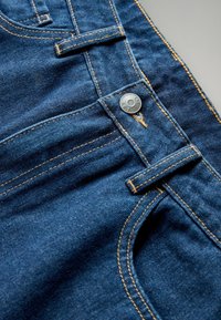 Tumši zilo džinsu bikses raksturo kontrastējošas dzeltenas šuvju līnijas, metāla poga un gluda tekstūra, kā arī klasiskā piecu kabatu dizains.