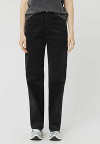 Pantalons en velours côtelé noir avec une coupe droite, présentant des poches avant, une fermeture à bouton et un tissu doux et texturé. Associés à des baskets grises.