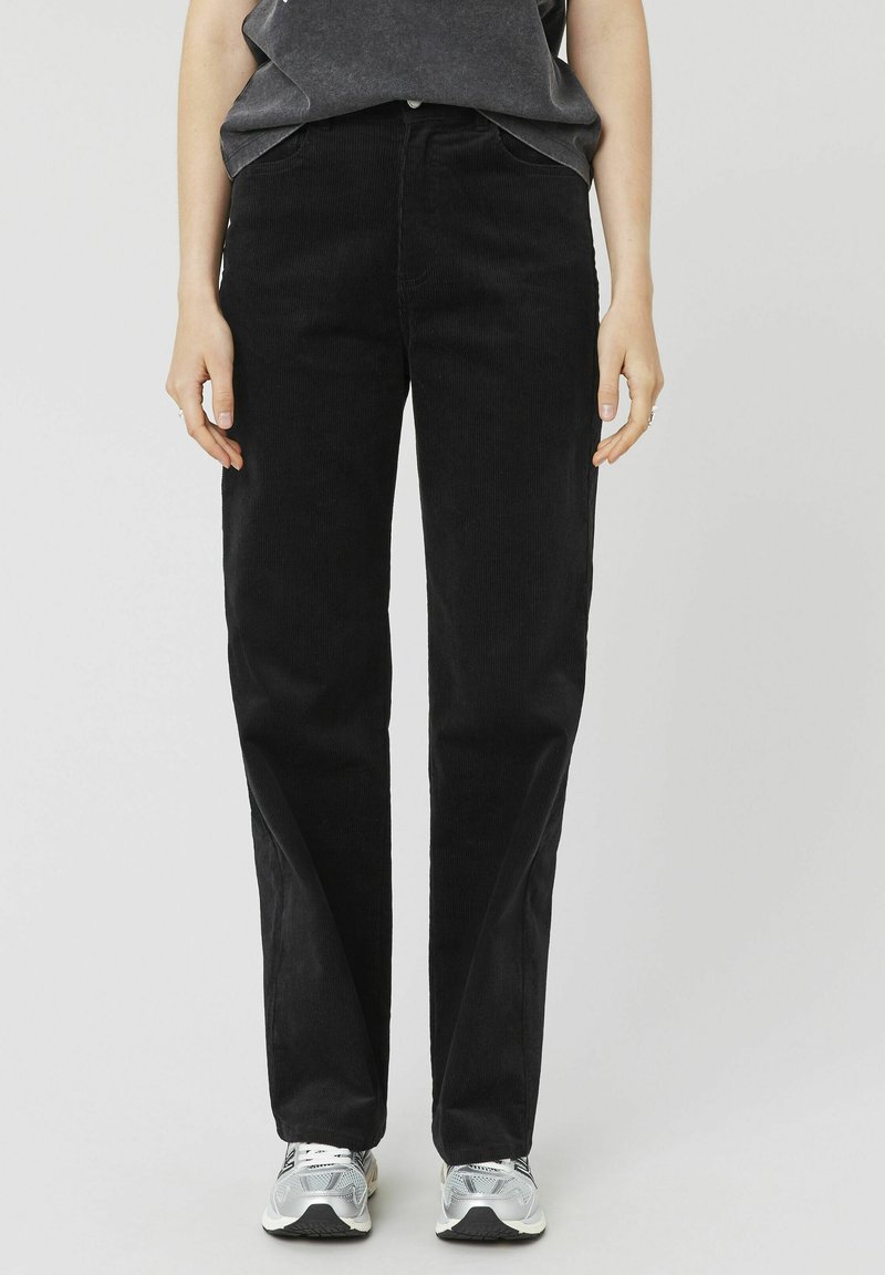 Pantalons en velours côtelé noir avec une coupe droite, présentant des poches avant, une fermeture à bouton et un tissu doux et texturé. Associés à des baskets grises.