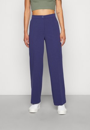 Persoon die marineblauwe broek met hoge taille en rechte pijpen draagt, met knoopsluiting en witte sneakers voor een effen achtergrond.