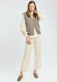 Vrouw in een crèmekleurige blouse en pantalon met een bruine gebuttonde gebreide vest en loafers met luipaardprint, staande met haar hand op haar nek.