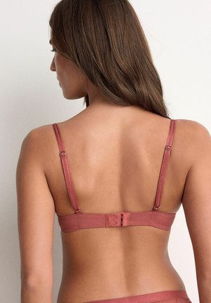 Sieviete ar gariem brūniem matiem, valkājot putekļrozā krāsas krūšturi, no aizmugures uz gaiša, vienkārša fona.