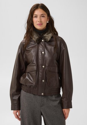 Femme portant une veste en cuir marron foncé avec col en fourrure et boutons-pression, associée à un pantalon gris taillé sur mesure, debout devant un fond uni.