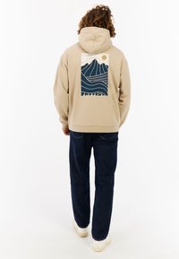 Beige hoodie met een grafische print van bergen en golven op de achterkant; heeft een trekkoord capuchon en ribgebreide zoom, gecombineerd met donkerblauwe spijkerbroek.