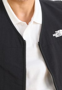 Chaleco negro con cremallera sobre una camisa tipo polo blanca. El chaleco presenta una textura suave y un logo en el área del pecho izquierdo.