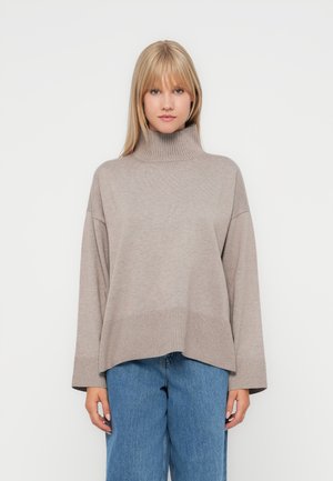 Pull col roulé beige clair avec une texture côtelée, des épaules tombantes et une coupe ample, associé à un jean en denim bleu décontracté.