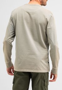Langärmliges Hemd aus hellem beigen Stoff mit rundem Ausschnitt und genähten Akzenten. Verfügt über einen geraden Saum und Handgelenkabschlüsse.