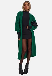 Cardigan verde a maglia, dolcevita nera a coste, gonna nera corta, collant trasparenti e stivaletti neri con tacco, accessorati con una pochette nera.