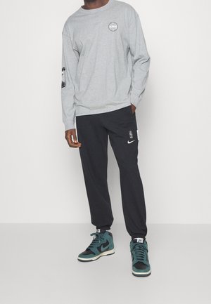 Personne portant un haut à manches longues gris clair, un pantalon de survêtement Nike NBA noir et des baskets montantes Nike turquoise et noires, debout sur fond blanc.