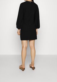 Robe noire à la silhouette ajustée, manches bouffantes et détail de dos en trou de serrure. Portée avec des sandales à talons en bois brun. Texture lisse.
