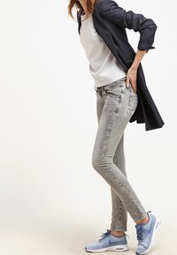 Jeans gris slim-fit, retroussés au niveau des chevilles, associés à un haut gris clair et un blazer marine, complétés par des baskets de sport bleues.