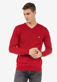 Roter Strickpullover mit V-Ausschnitt, gerippten Bündchen und Saum, der ein kleines schwarz-weißes Logo auf der linken Brust hat.