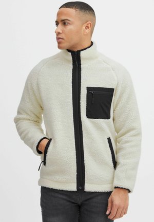 Fleece jacket in off-white with textured exterior, black zip pockets, high collar, and front zipper. Casual design with contrasting accents.
