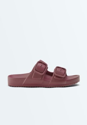 Einzelne mauvefarbene Sandale mit Keilabsatz, ausgestattet mit zwei großen, verstellbaren Schnallenriemen und strukturierter Sohle, seitlich auf weißem Hintergrund dargestellt.