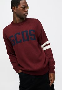 Bordowa dzianinowa bluza z dużym granatowym logo „GCOS” i białymi paskami na rękawach. Żebrowany dekolt i mankiety.