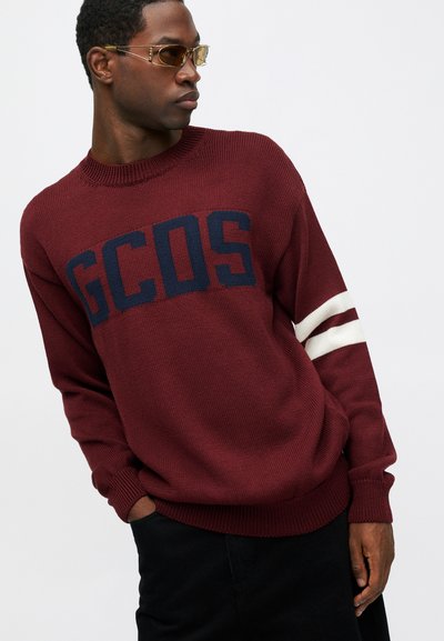 Burgunderfarget strikkegenser med en stor marineblå "GCOS"-logo og hvite striper på ermene. Ribbestrikket halslinning og mansjetter.