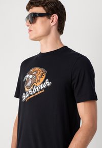Tricou din bumbac negru cu un grafic al unui tigru miorlăind în nuanțe de portocaliu și maro, având textul "Barbour International" dedesubt în alb și portocaliu.