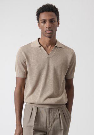 Joven con cabello corto y rizado que lleva una camisa polo de punto beige de manga corta y pantalones plisados beige, de pie frente a un fondo liso.