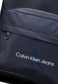 Borsa in tessuto navy con tasca anteriore con zip, logo "Calvin Klein Jeans" in bianco e blu scuro, con una superficie liscia e texture.