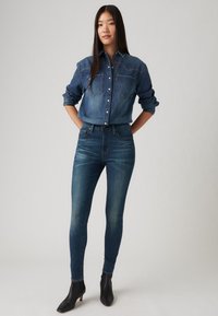 Žena stojaca s rukami za chrbtom, oblečená v džínsovej košeli, tmavomodrých obtiahnutých nohaviciach a čiernych členkových čižmách na jednorazovom pozadí.