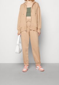 Person som bär en beige huvtröja och matchande byxor, en grön kort topp, rosa och vita sneakers, håller en vit handväska vid sidan.