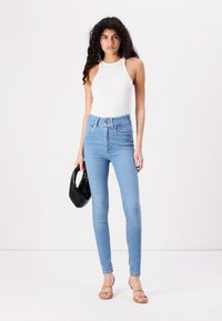 Tynne blå jeans laget av denim, med høy midje, to frontlommer og knappeåpning. Kombinert med en hvit ermeløs topp.