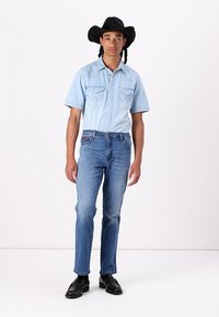 Lys blå denim skjorte med korte ærmer, lommer og stingdetaljer, parret med medium blå jeans og sorte sko, båret med en sort cowboyhat.