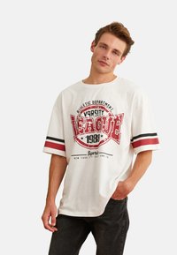 Camiseta blanca de gran tamaño con estampado gráfico en rojo, texto histórico y mangas a rayas negro/rojo. La tela parece suave; ajuste casual.