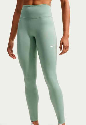 Kobieta nosząca wysokowalne legginsy Nike w kolorze szałwiowym z białym logo Swoosh na prawej uda, stojąca z ramionami swobodnie opuszczonymi wzdłuż ciała.