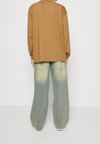 Ruskea pitkähihainen paita rennolla leikkauksella yhdistettynä leveälahkeisiin farkkuihin, joissa on haalistunut sininen väri ja kaksi takataskua.