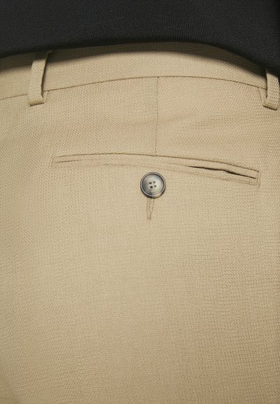 Beige nadrág, texturált anyagból, egy hátsó zsebből és egy szürke gombbal záródó zsebbel.