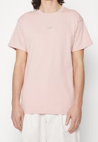 Ljusrosa bomullströja med korta ärmar, rundad halsringning och diskret vit Nike-logga på bröstet. Slät struktur och avslappnad passform.