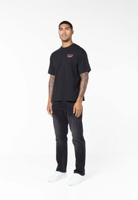 Muž s tetovanými pažemi, oblečený v černém tričku, černých džínách a bílých teniskách stojí na bílém pozadí.