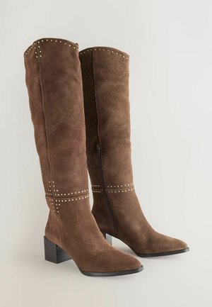 Bruna mockaskinn knähöga stövlar med spetsig tå och klack i blockstil. Funktioner inkluderar silvernitar längs toppen och horisontell söm. Sidodragkedja.