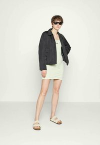 Jaqueta preta cropped sobre um vestido verde claro, usada com sandálias creme. A modelo usa óculos escuros escuros e tem cabelo curto e encaracolado.