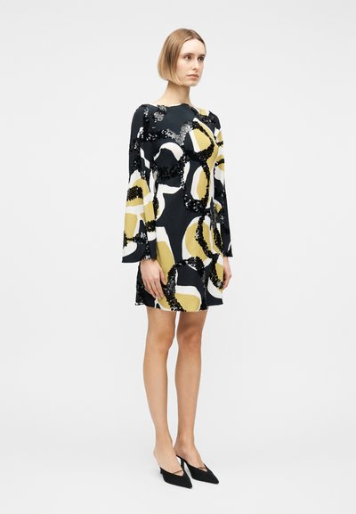 Γυναίκα που φοράει μακρυμάνικο φορεμα μέχρι το γόνατο με αφηρημένα μοτίβα σε μαύρο, άσπρο και κίτρινο, σε συνδυασμό με μαύρες μυτερές γόβες.