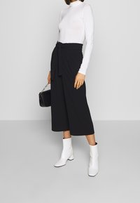 Culottes noirs à jambes larges avec une taille nouée, associées à un turtleneck fitted à manches longues blanc et des bottines blanches. Sac à main noir texturé.