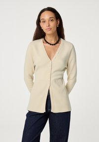 Cream cardigan with a v-neck, front button closure, ribbed texture, and long sleeves, paired with dark denim pants.