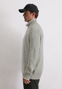 Sweater en tricot texturé gris clair avec un col montant et une fermeture éclair. Présente des poignets et une base côtelés, porté avec une casquette noire.