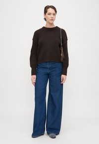 Pulover gros tricotat de culoare maro, cu guler rotund, purtat împreună cu blugi din denim albastru, cu picior larg. Modelul ține o geantă cu detalii cu ciucure.