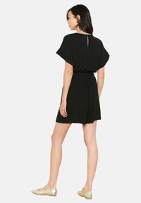 Czarny kombinezon z krótkim rękawem, ze zwężoną talią i otworem w kształcie klucza z tyłu, wykonany z miękkiego, teksturowanego materiału. W połączeniu ze złotymi balerinami.