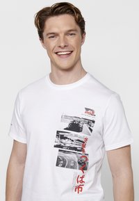 Balta medvilninė marškinėlių suapvalintu kaklu, trumpomis rankovėmis, kurių dizainą sudaro juodieji ir raudonieji automobiliai bei tekstas.